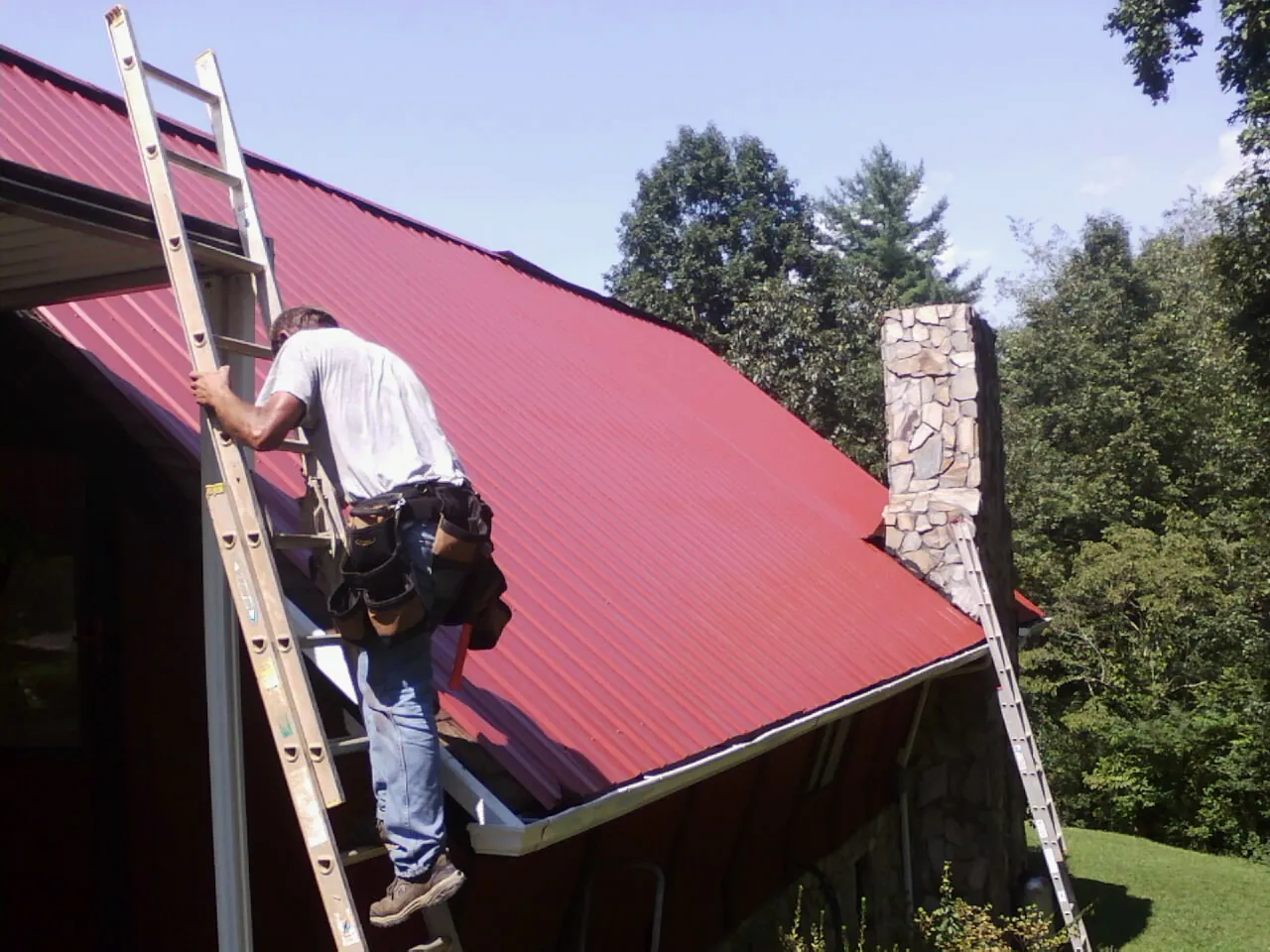 Roofer on a ladder beside a red metal roof and stone chimney — Soffit & Fascia Repair by Roofing Brothers in Brownsdale